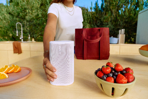 Sonos Era 100 speaker in white, shown plugged into a wall with a woman's finger touching the top of the speaker.