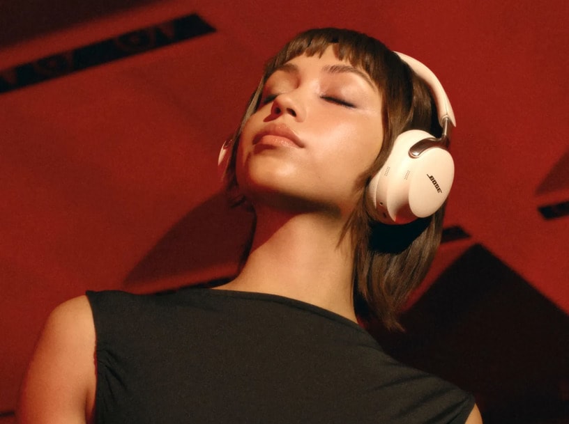 Person wearing white Bose QuietComfort Ultra Headphones 2nd Gen with eyes closed in an upward gaze, photographed from below against a red background with dramatic shadows, wearing a black sleeveless top.