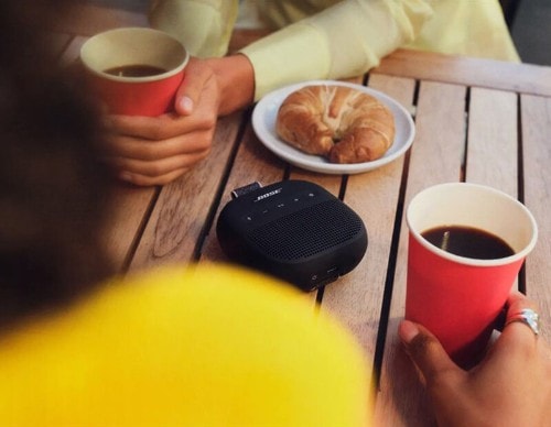 Two people at outdoor wooden table with red coffee cups and pastry, Bose SoundLink Micro black speaker placed on table between them.