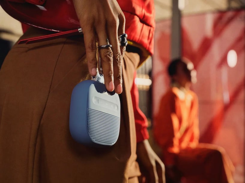 Bose SoundLink Micro Portable Speaker (2nd Gen) in Blue Dusk clipped to a person's pants with a carabiner, shown in a casual outdoor lifestyle setting.