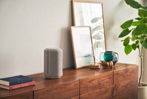 Sony light gray speaker on wooden sideboard with framed art, decorative glass vase, book, and plant in modern minimalist interior