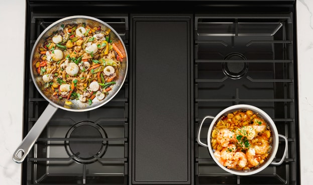 Overhead view of Samsung gas range cooktop with five burners showing two pans with food cooking, demonstrating burner configuration and cooking capacity.