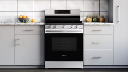 Front view of Samsung NX60A6111SS stainless steel gas range with black oven door and sleek control panel, installed in kitchen with light gray cabinets and white tile.