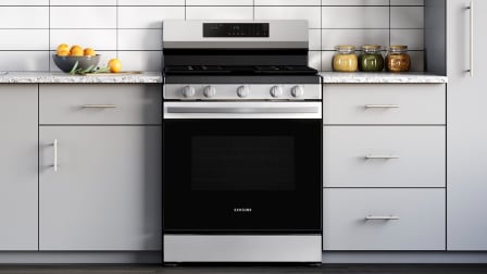 Front view of Samsung NX60A6311SS stainless steel gas range with black oven door and control panel, installed in kitchen with light gray cabinets and white tile backsplash