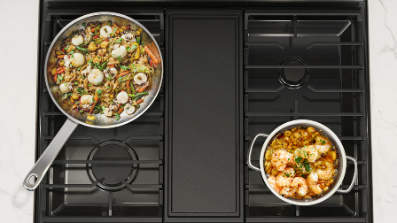 Overhead view of Samsung gas range black cooktop with continuous cast-iron grates supporting two cooking pans with food, demonstrating five-burner configuration.