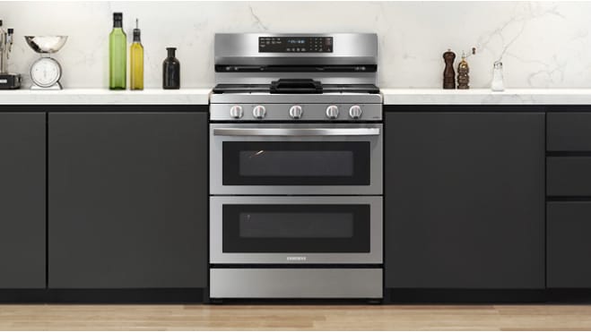 Samsung NX60A6751SS stainless steel gas double oven range installed in modern kitchen with black cabinets, white marble backsplash, and white countertop.