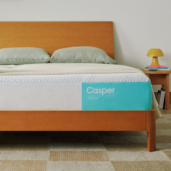 Casper Snow mattress on wood platform bed frame with white quilted cover and turquoise Casper branding, gray pillows, cream bedding, wood nightstand with yellow lamp and books.