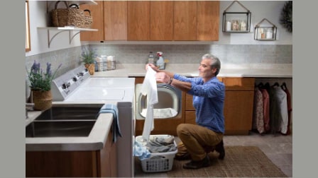 Man kneeling beside laundry basket loading white top-load Speed Queen washer in modern laundry room with wood cabinets, white countertops, and gray tile backsplash.
