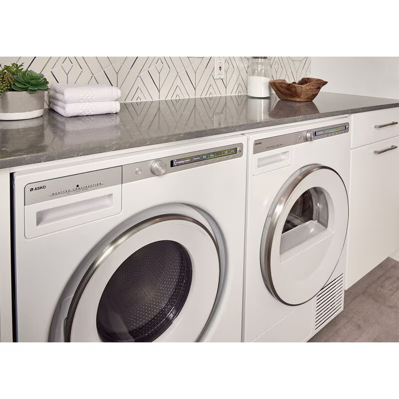 Asko front load washer and dryer set in white finish installed in modern laundry room with gray countertop, geometric tile backsplash, folded towels, and decorative accessories creating elegant functional space.