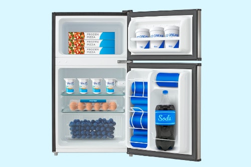 Interior view of compact refrigerator showing top freezer with frozen foods, main compartment with wire shelf, crisper drawer with blueberries, and door shelves holding beverages.