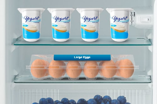 Interior view of refrigerator showing two glass shelves with yogurt cups on top shelf and egg carton on middle shelf, demonstrating organized storage space.