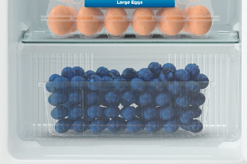 View of refrigerator crisper drawer filled with fresh blueberries below glass shelf holding egg carton, showing fresh food storage and humidity control features.