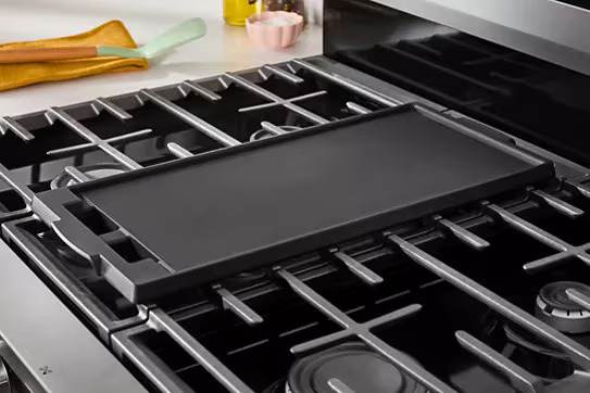 Close-up of Whirlpool WFGS5030RB gas range cooktop showing cast iron grates with removable center griddle insert positioned over center burner, black porcelain cooktop surface with yellow and pink kitchen accessories in background.