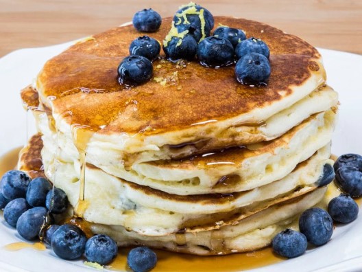 Stack of four blueberry pancakes with blueberries, lemon zest, and syrup on top