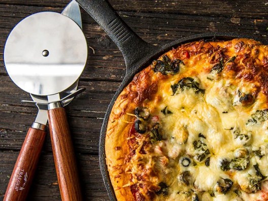 Overhead shot of deep dish pizza in skillet on wooden table, with pizza cutter and server next to it