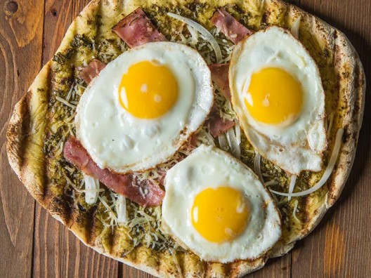 Overhead shot of breakfast pizza on wooden table, topped with three sunny-side-up eggs