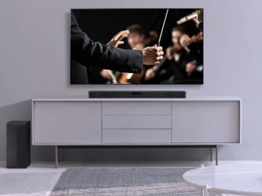 Large TV in Gray Minimalist Living Room Outfitted with Soundbar and Subwoofer, and Displaying Image of Conductor's Hands Holding Baton in Front of Orchestra