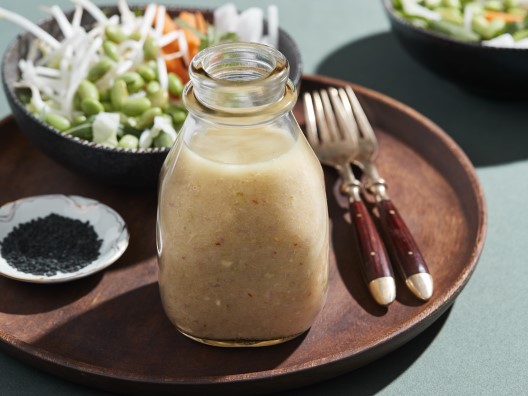 Small Bottle of Salad Dressing with Forks, Sesame Seeds, and Salads in Background