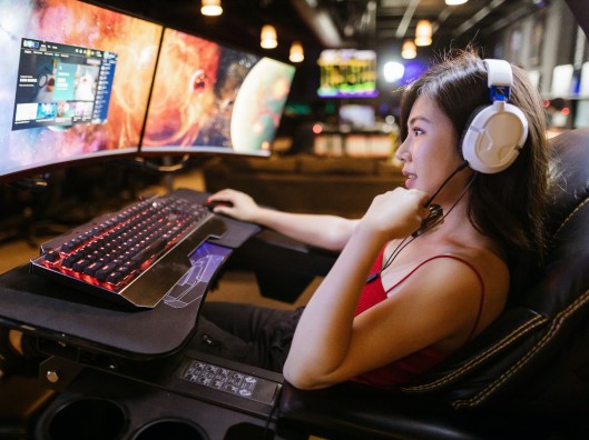 Asian Woman Wearing Headset and Sitting in Gaming Chair at Dual-Monitor Gaming PC