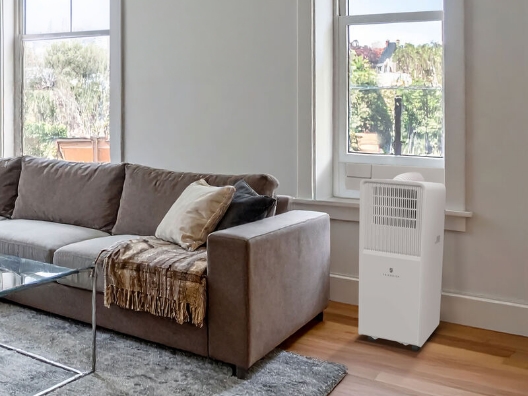 A portable air conditioner sitting next to a couch and in front of a window.