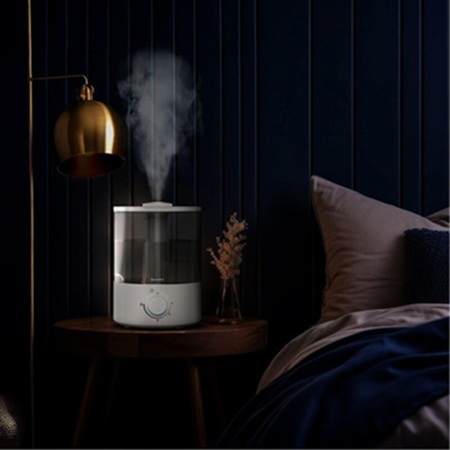 An air humidifier sitting on a bedside table next to a bed and under a lamp.