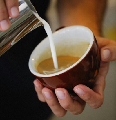 Person pouring steamed milk into a cappuccino mug containing espresso