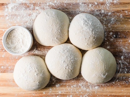 Five balls of pizza dough next to a small bowl of flour on a wooden work surface