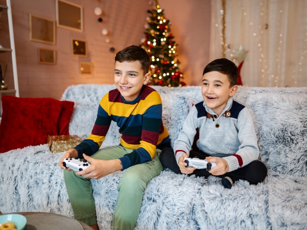 Two young boys sitting on a couch and playing video games with a Christmas tree in the background.