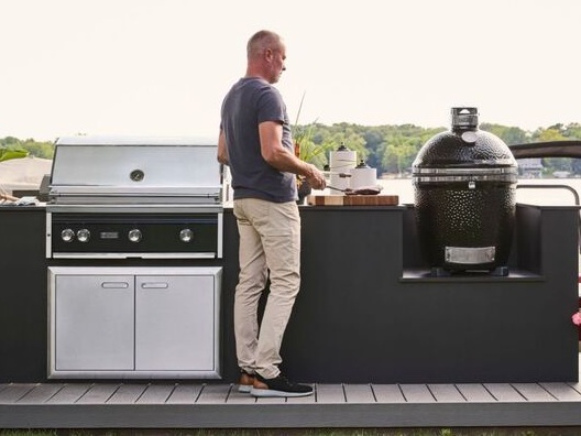 A man standing in front of an outdoor kitchen with a gas grill and a charcoal grill.