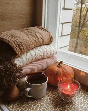Blankets stacked near a pumpkin, a mug, a candle, and a plant.