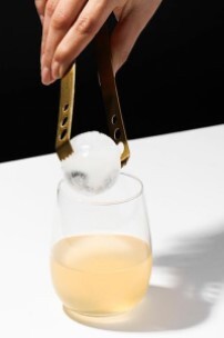 A hand with a pair of metal tongs placing an ice sphere in a glass