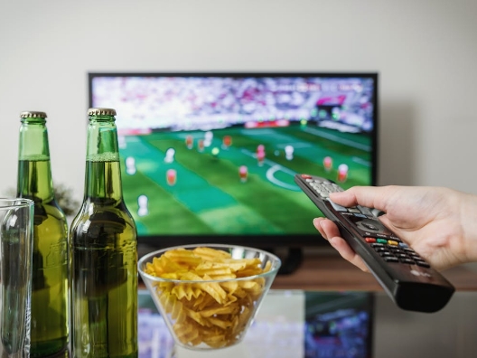 A person pointing a remote at a TV over a bowl of chips and drinks.