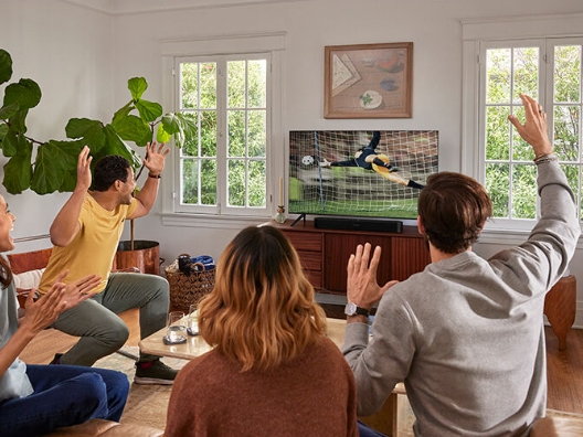 A group of people excitedly watching soccer on a TV with a soundbar placed in front of it.