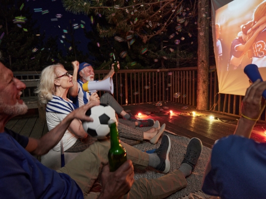 A group of older people outside on a terrace watching a soccer match on a projector and drinking beverages.