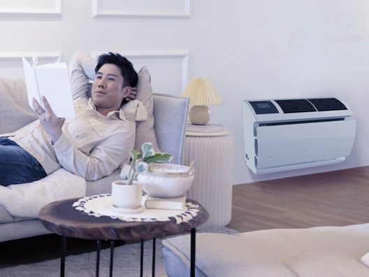 A man laying on a couch reading next to a wall air conditioner.