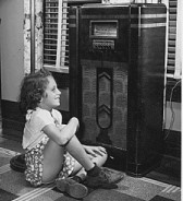 Black and white 1940s photo of a girl sitting on the floor in front of a vintage radio