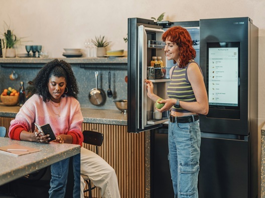 Two women talking in front of an open smart refrigerator.