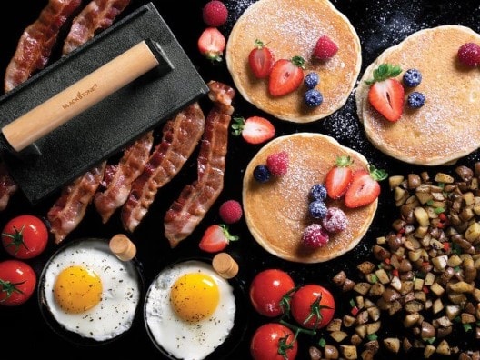 Overhead view of Blackstone griddle with pancakes, eggs, home fries, and bacon under a griddle press