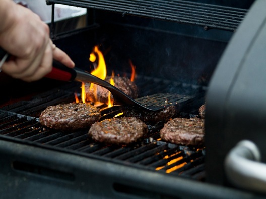 A person pressing a spatula onto a hamburger patty on a flaming grill.