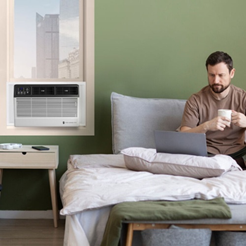 A man sitting on a bed looking at a laptop next to a window air conditioner.