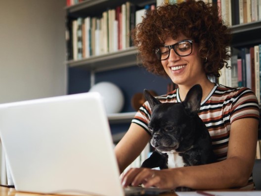 A woman with curly hair typing on a laptop while a small dog sits in her lap